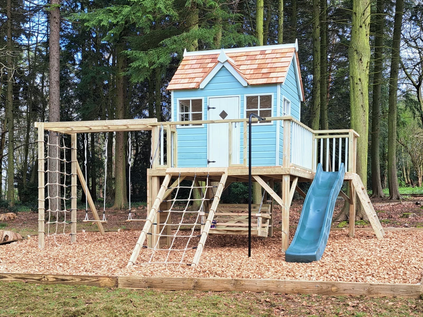 An Otter Playhouse: the painted Otter cottage with cedar shingle roof on its full play deck, with wave slide, monkey bars and swings