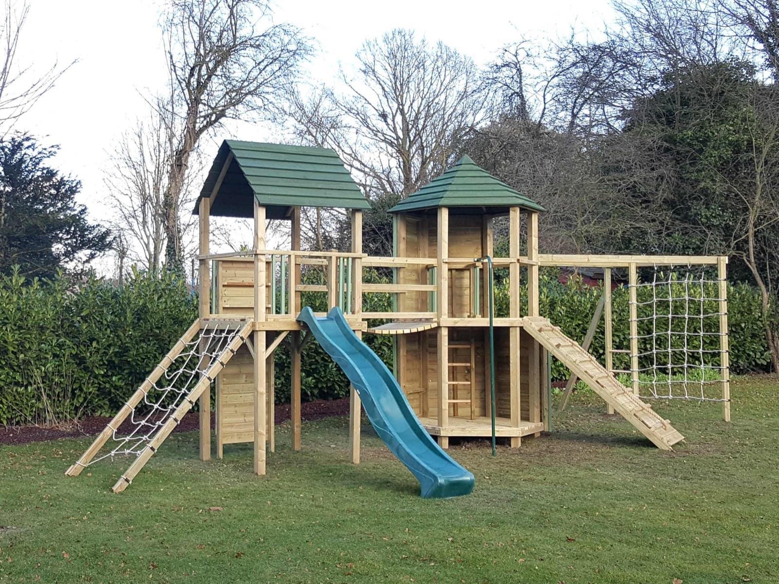 A Treetops Hex Multi-Tower: a green-roofed hexagonal tower decked over two levels beside a square tower, with a wave slide, sloping scramble net and climbing wall