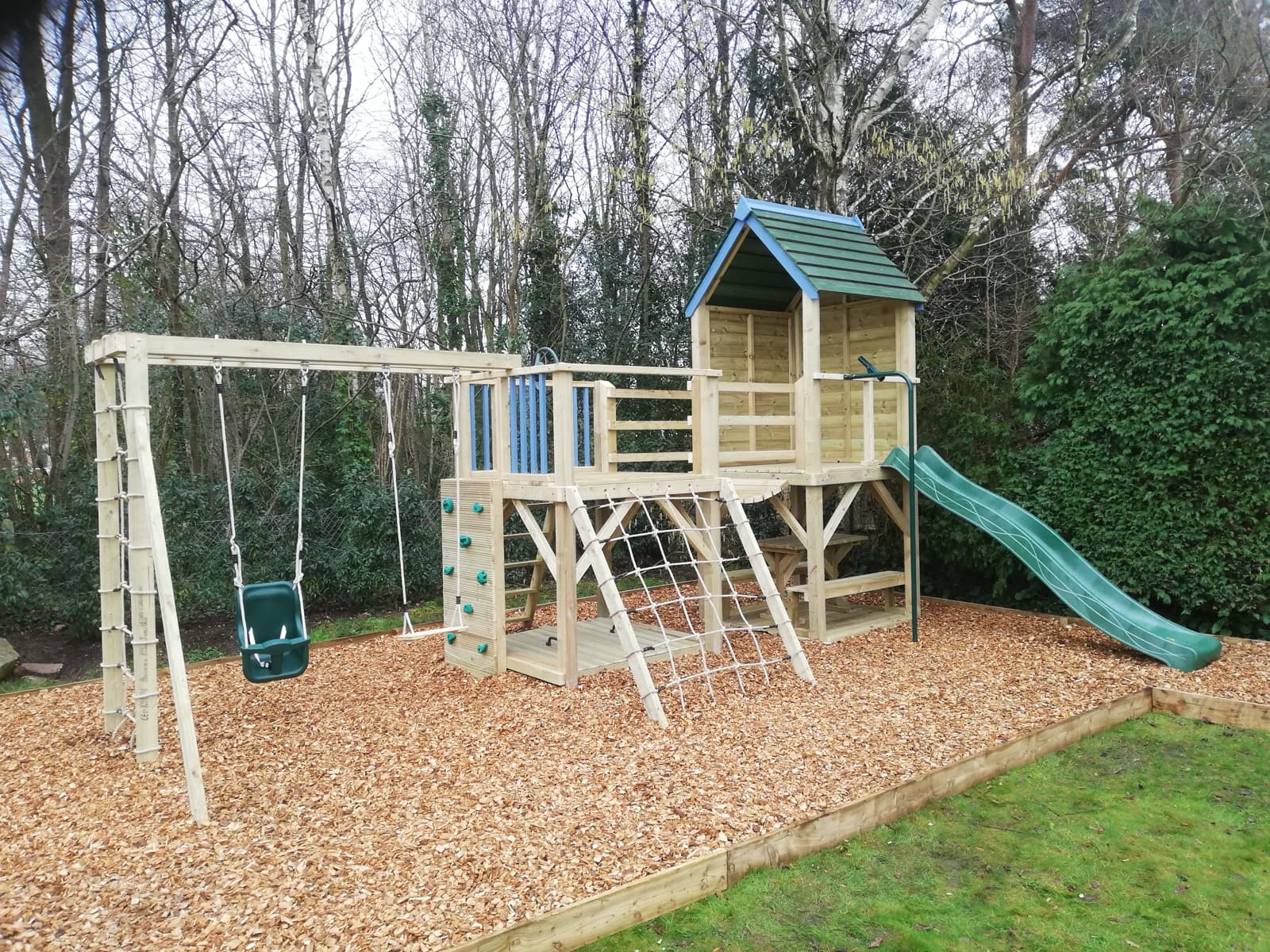 A Treetops Multi-Tower: two timber towers joined by a chain-link clatter bridge, with a wave slide and scramble nets
