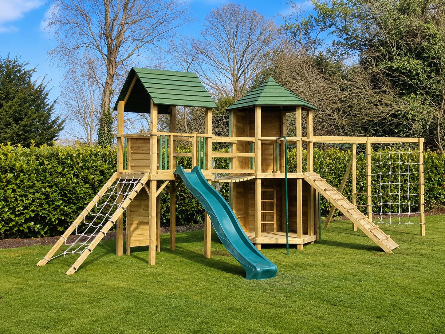 A Treetops Hex Multi-Tower climbing frame with green-roofed towers, wave slide, and scramble nets in a hedged garden