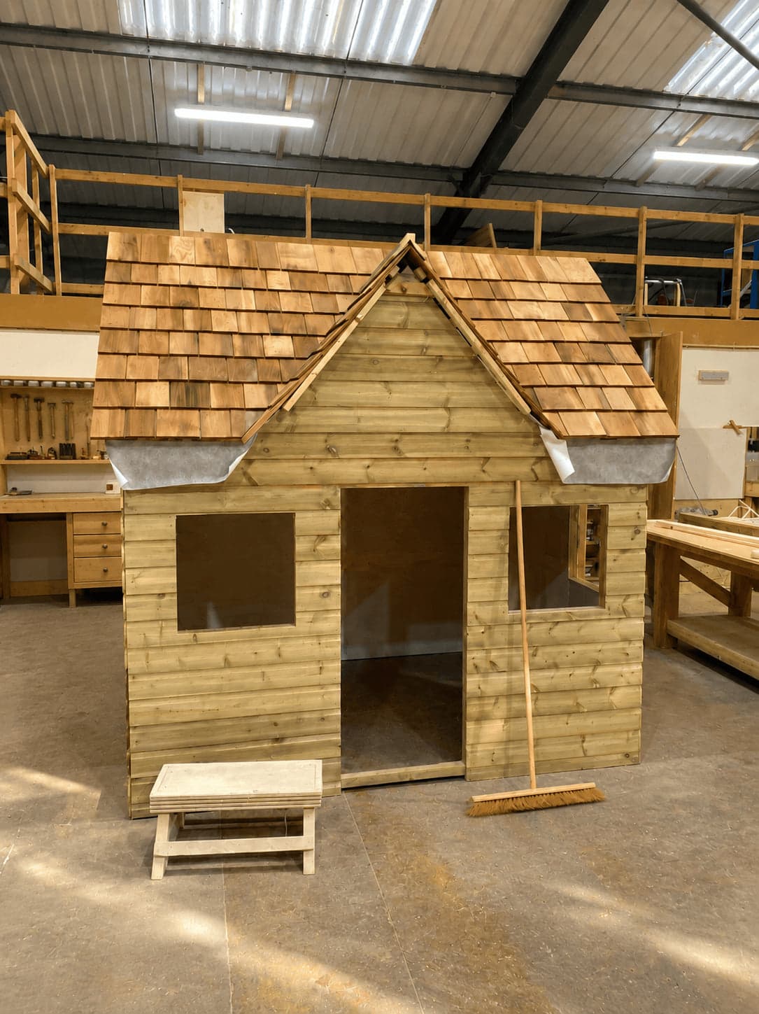 A playhouse standing complete on the workshop floor, cedar shingles laid and membrane still visible at the eaves