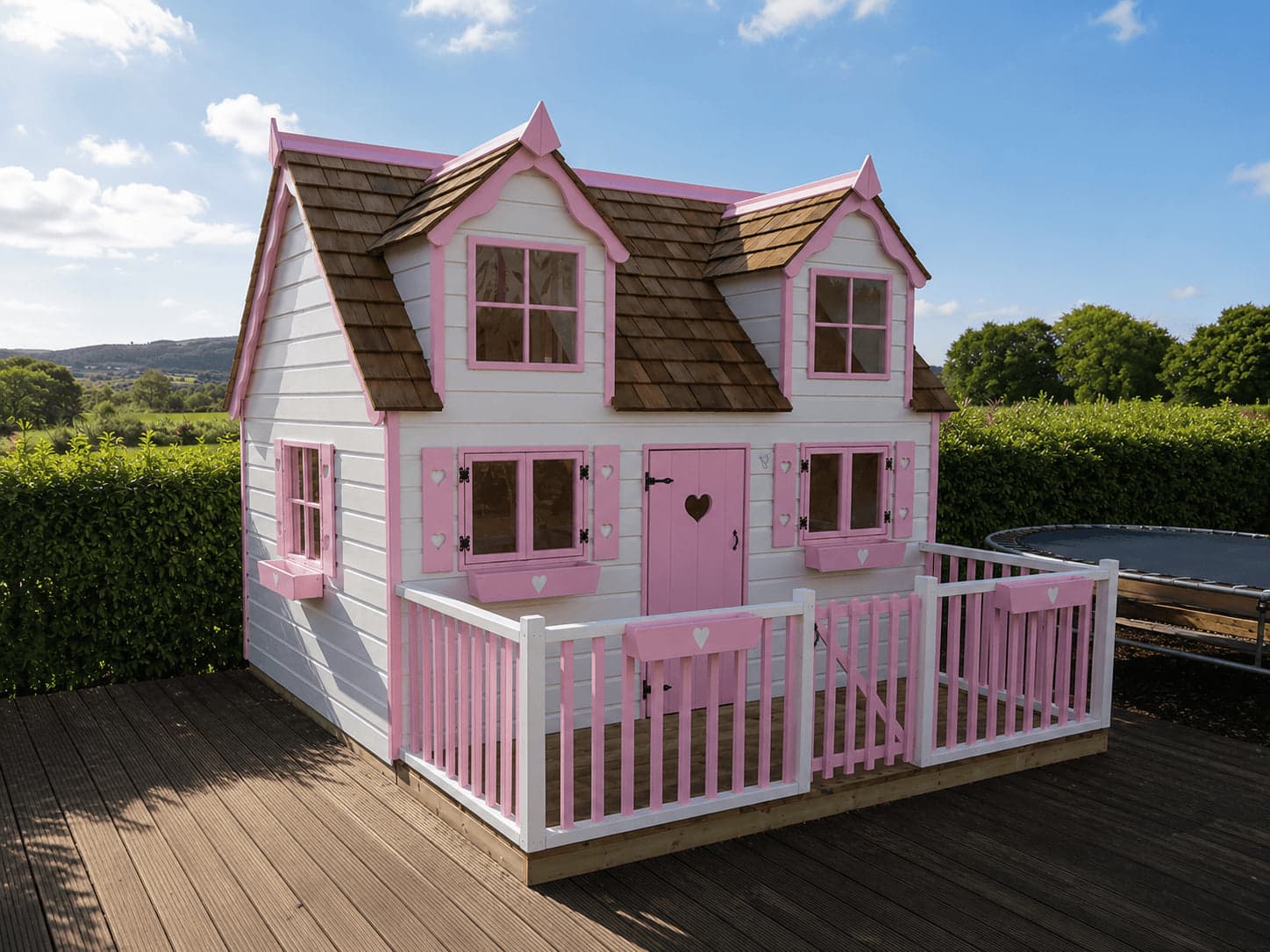 A white and pink two-storey playhouse with a cedar shingle roof, dormer windows, heart cut-outs and a gated picket deck