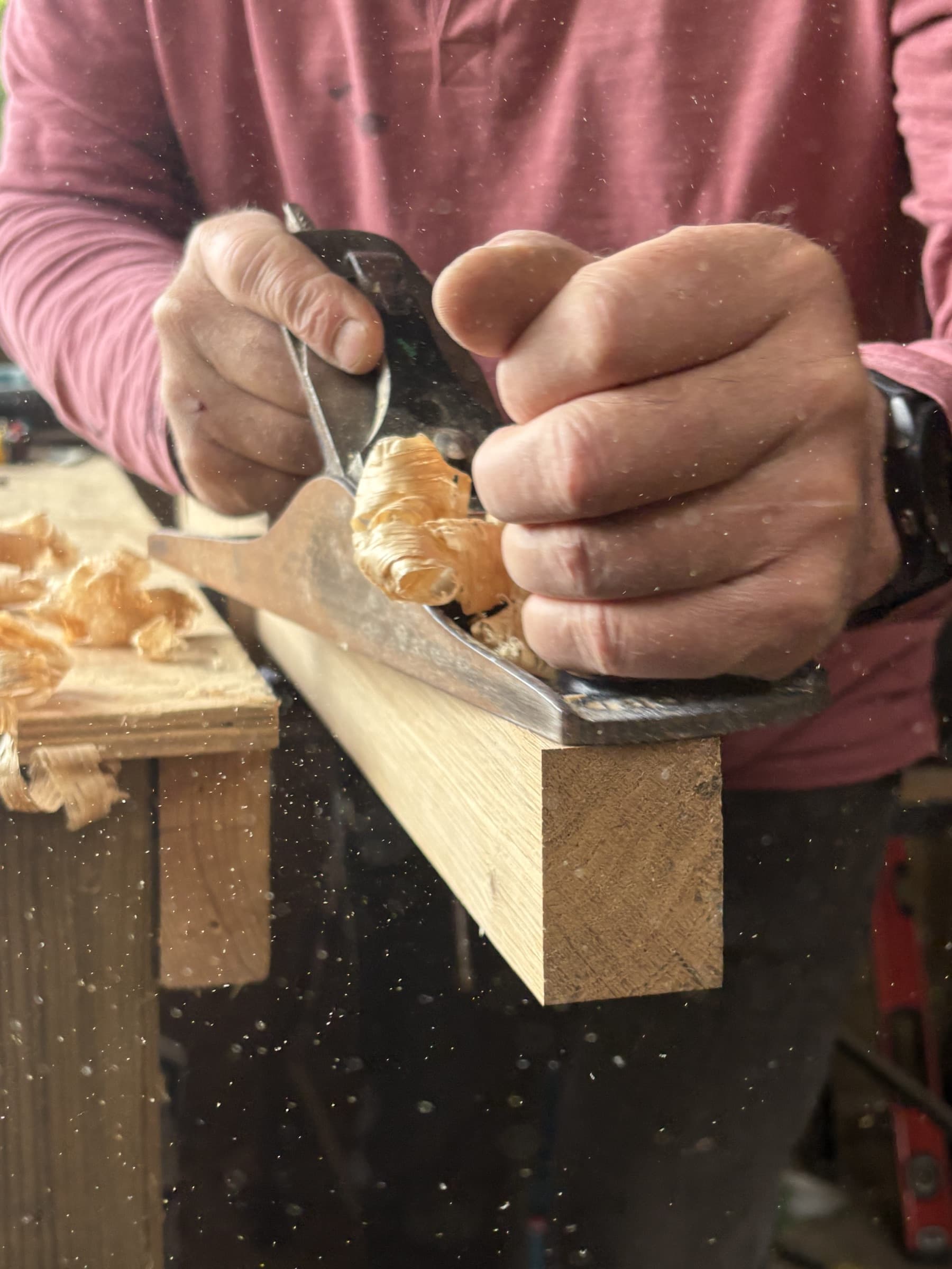 Hands guiding a plane along timber in the BESPOKE workshop, shavings curling from the blade