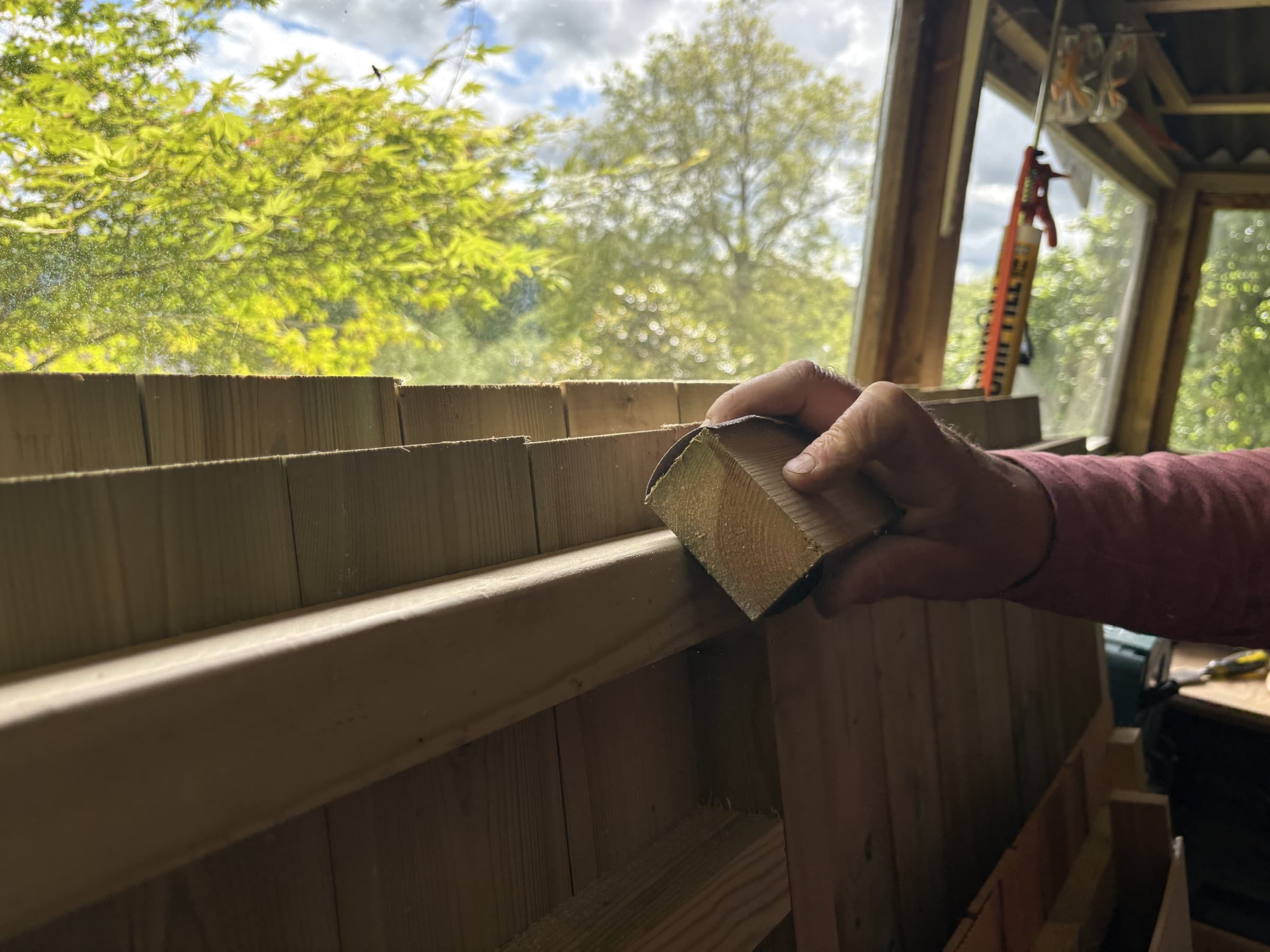 A craftsman fitting timber against fresh shingle courses at the workshop window, garden light beyond